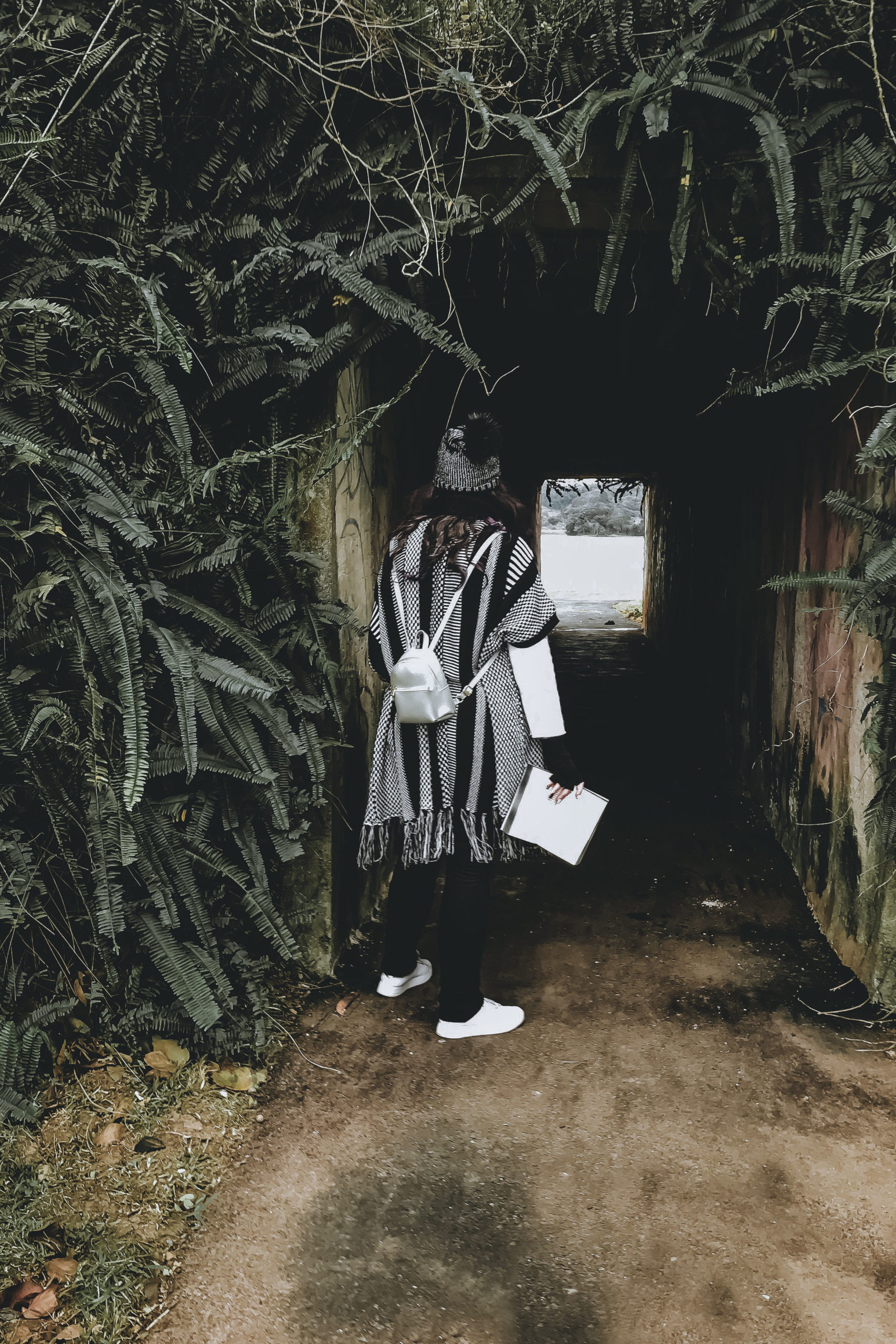 foto de uma mulher de costas, olhando para um túnel que passa entre samambaias. A mulher está vestida com roupas de inverno e segura um livro aberto na mão.